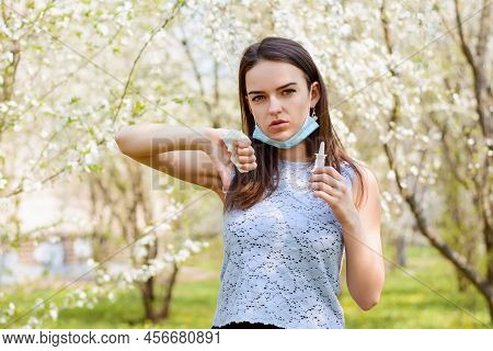 Young Girl In Medical Mask Showing Thumbs Down Standing In Blooming Garden And Holding Spray For Run
