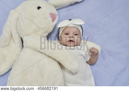 White Dreesed Newborn Baby With Her White Bunny Toy. Cute Mixed Race Baby Girl Looking Into The Came