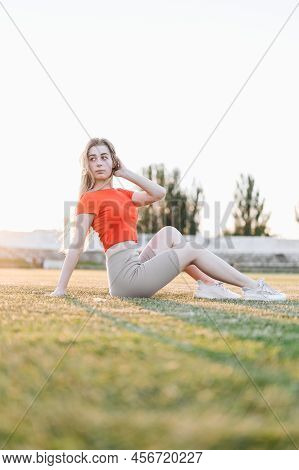 Athletic Girl Sits Between Exercises On Green Grass With Her Knees Bent And Looks To The Side. Woman