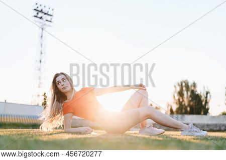 Athletic Girl Lies On The Green Grass With Her Knee Bent And Looks To The Side. Girl In Sportswear I