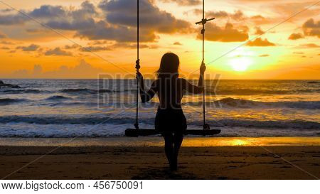Young Woman Swinging Sitting On Wooden Swing With Ropes On Sandy Beach, Sea Sunset Background. Girl 