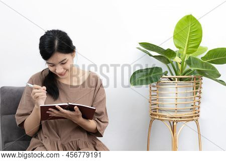 Asian woman sits in her living room writing a diary or notebook on the carpet, Weekends or holidays 