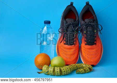 Orange Sneakers, Measuring Tape, Water Botlle And Fruits Apples And Oranges On A Blue Background. We
