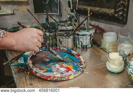 The Artist Holds A Brush In His Hand Near A Palette With Bright Colors In An Art Workshop