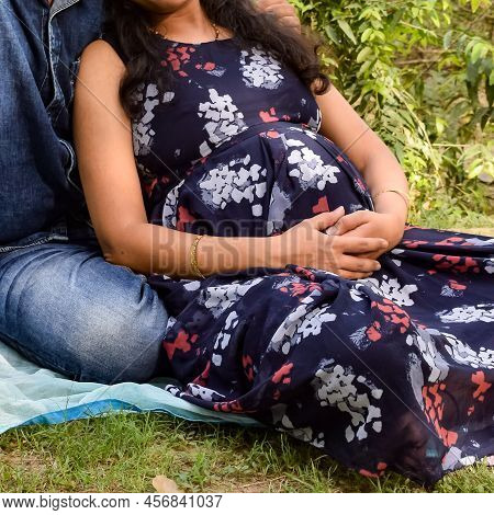 Indian Couple Posing For Maternity Baby Shoot. The Couple Is Posing In A Lawn With Green Grass And T