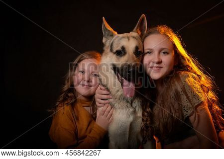 Two Cheerful Joyfull Sisters Having Fun Together And With Big German Shepherd Dog In Dark Studio Wit