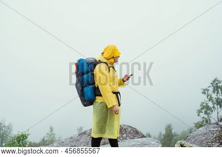 A Tourist On A Hike With A Backpack On His Back Stands On A Rock On A Background Of Clouds With A Sm