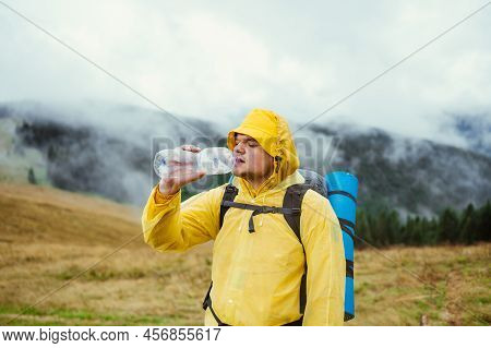 Hiker In A Yellow Raincoat On A Hike In The Rain Drinks Water From A Bottle On A Background Of Beaut