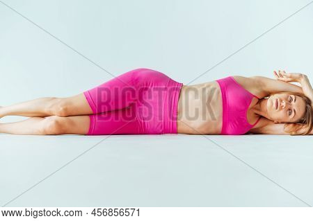 Athletic Woman In Pink Clothes Lying On The Ground On A White Background And Looking At The Camera. 
