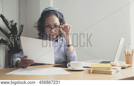 Seriously Senior Woman Tworking At Home, Looking At Laptop And Waving Paper Correspondence, Sitting 