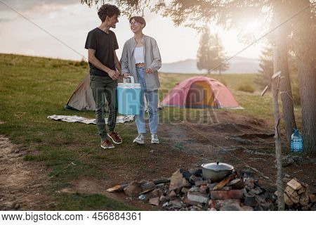 Joyful Young Man And Woman Standing At Camping Stop With Portative Fridge And Laughing.