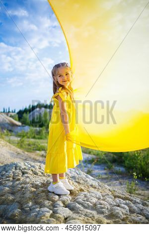 A Small 6-7 Years Old Ukrainian Girl In A Yellow Dress With Wings In Yellow Fabric By The Lake. Blue