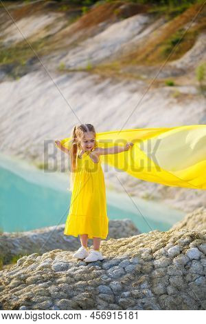 A Small 6-7 Years Old Ukrainian Girl In A Yellow Dress With Wings In Yellow Fabric By The Lake. Blue