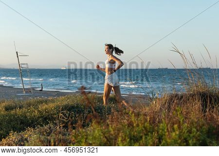Side View Of Young Woman In Sports Bra And Wireless Earphone Jogging On Grass Near Sea Shore.