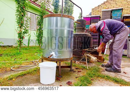 Man Throws Dry Branches Into The Firebox Of A Homemade Distillery Making Moonshine Schnapps, Alcohol