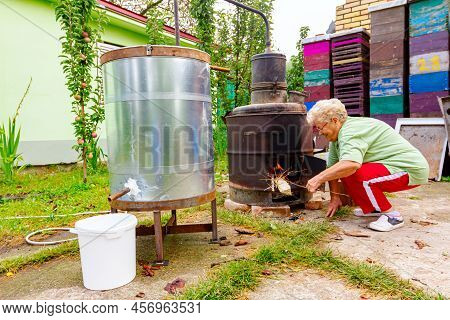 Grandma Throws Dry Branches Into The Firebox Of A Homemade Distillery Making Moonshine Schnapps, Alc