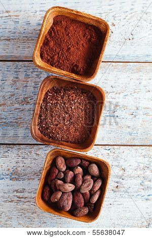 cocoa beans, powder and grated chocolate in wooden bowls, white wood backdrop