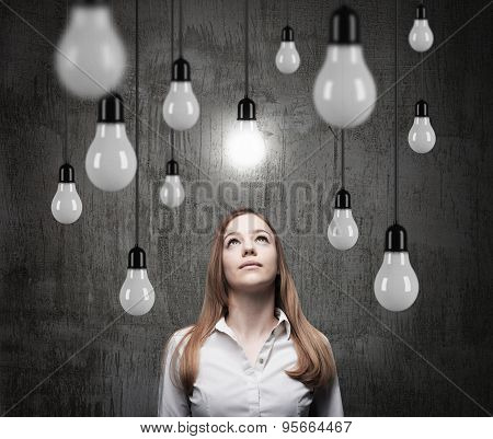 Charming Lady Is Looking Upward At The Hanging Light Bulbs. A Concept Of Searching New Ideas. Dark C