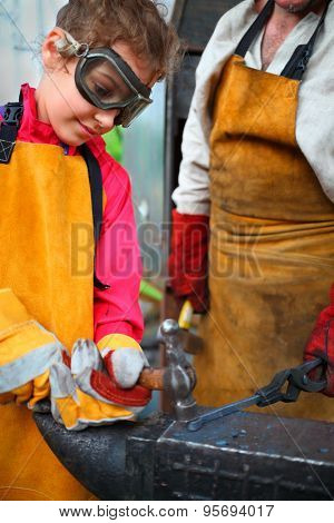 Girl in goggles and yellow apron with  hammer hitting anvil in smithy