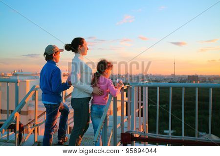 Mother with son and daughter standing on roof of multistory house and looking into distance