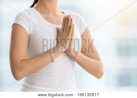 Young yogi woman practicing yoga, making namaste gesture, working out, wearing white t-shirt, chain pearl wrist bracelets, indoor, meditation session near the window. Midsection close up