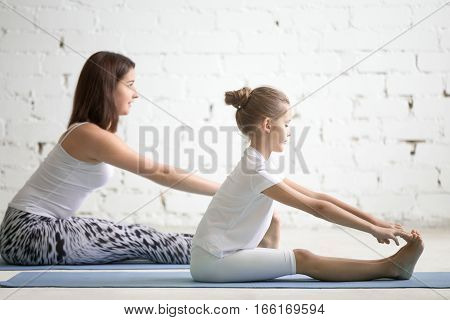 Kids yoga female teacher training a girl child, two people practicing yoga, sitting in paschimottanasana exercise, seated forward bend pose, working out wearing sportswear, indoor, white studio