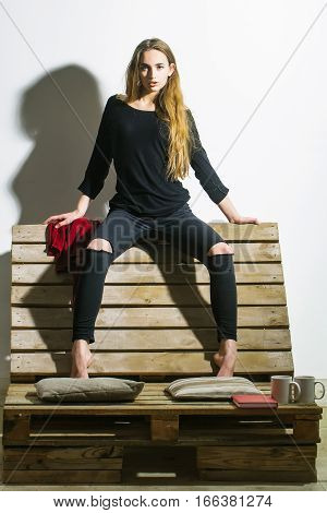 Pretty girl or beautiful woman cute sexy fashion model with long hair in casual clothes sits on backrest of wooden pallet sofa on white wall