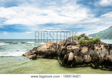 Rock formation in the middle of the sea. Waves hitting the rocks and a green mountain on the background on a very tranquil scene. Praia do Matadeiro Florianopolis Brazil.