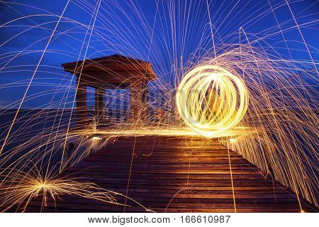 Hot Golden Sparks Flying from Man Spinning Burning Steel Wool on Wooden Bridge Extended into the Sea. Long Exposure Photography using Steel Wool Burning.