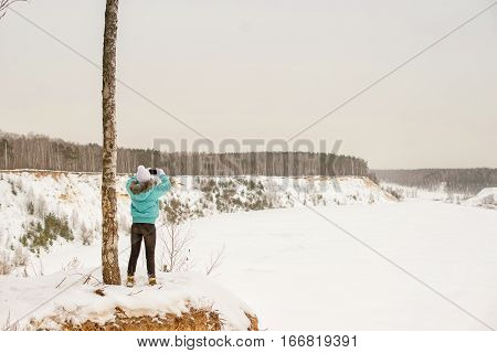 Girl in bright clothes on the edge of the cliff on the background of the winter forest in winter. In the hands of the phone take pictures on the phone