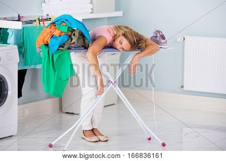 Young Tired Woman Sleeping On Ironing Board Next To Pile Of Clothes At Home