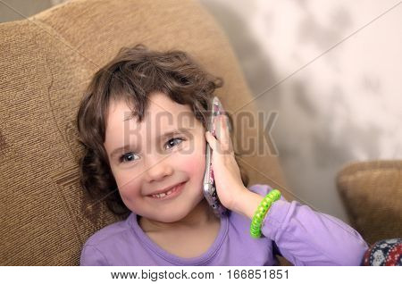 Cute little girl is talking on the mobile phone and smiling while sitting on couch at home. Small girl with smartphone. Girl talking on smartphone.  Little girl using smartphone. Smartphone in hand.