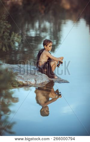 Caveman Boy Sitting On The Rock Near River Or Lake And Looking Away. Evolution Survival Concept. Cre