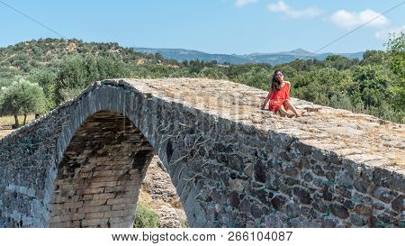 Girl In Red Dress Sitting On Ancient Bridge With Mountains In Background