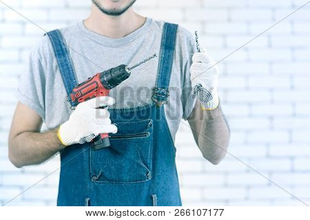 Young Asian Man Holding Power Drill Standing In Front Of White Brick Wall, Smiling And Looking At Ca