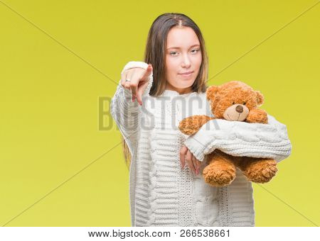 Young caucasian woman holding teddy bear over isolated background pointing with finger to the camera and to you, hand sign, positive and confident gesture from the front