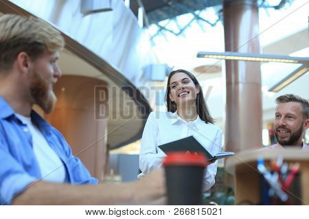 Group Of Young People In Casual Wear Sitting At The Office Desk And Discussing Something While Looki