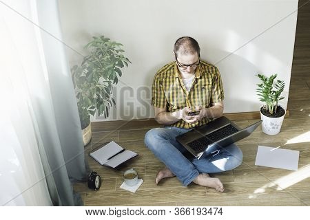 Young Man Sitting On The Floor And Working At Home On A Computer With The Phone, Papers And A Cup Of