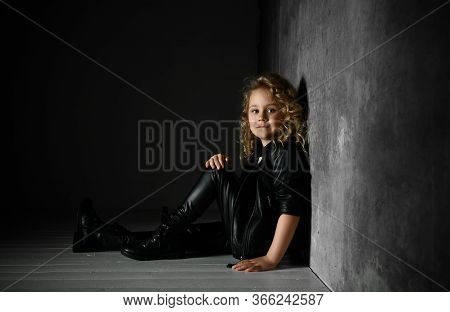 Adorable Little Girl With Curly Golden Hair Dressed In Black Sitting In Spotlight On The Floor Close