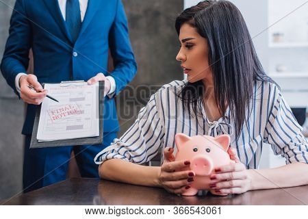 Woman With Piggy Bank At Table Near Collector Pointing With Pen At Documents With Final Notice Lette