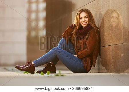 Happy young fashion woman sitting on street sidewalk  Stylish female model in brown leather fringe suede jacket and dark blue jeans