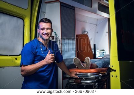 A Young Paramedic In A Blue Uniform Taking Away His Patient By An Ambulance, Smiling And Showing Thu