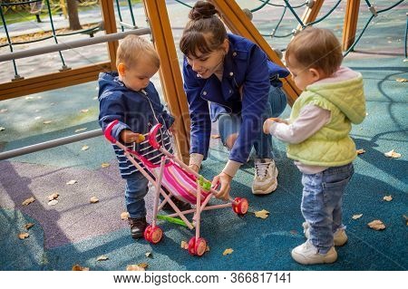 Young Beautiful Mother In A Blue Jacket An Autumn Park Plays Toy Stoller With Her Little Cute Toddle