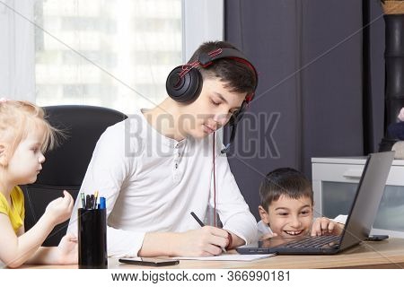Teenager Looks At A Laptop And Writes Down A Task And Children Interfere With Learning