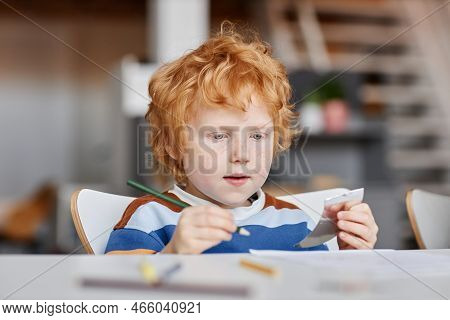 Cute Diligent Gingerhaired Learner Of Nursery School With Crayon Of Dark Green Color Drawing By Desk