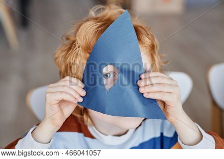 Eye Of Cute Gingerhaired Little Learner Of Nursery School Looking At Camera Through Hole In Dark Blu