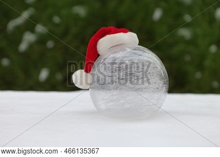 White Round Glass Ball On Snow With Christmas Hat. Winter World Near Christmas Tree. Snowy Cold Outd