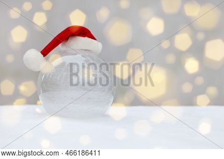 Transparent Glass Ball On Snow With Christmas Hat. Snow And Bokeh Flare On Santa Claus Hat. Snowy Ba