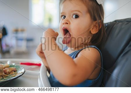 Happy Baby Girl Eating With Pleasure Food By Hand, Sitting In High Chair At Table With Plate. Baby-l