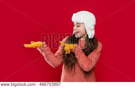 Teen Child In Earflap Hat Presenting Product Isolated On Red, Copy Space. Teen Child Wear Earflap